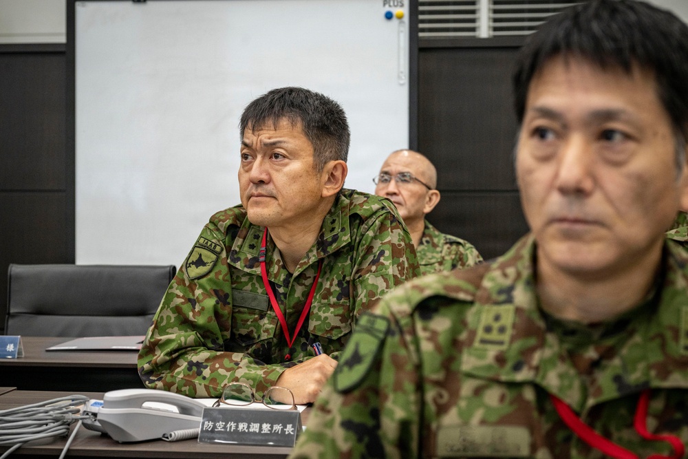 Three military personnel in Japanese Ground Self-Defense Force (JGSDF) camouflage uniforms are seated at a conference table in what appears to be a briefing or command room. A nameplate in Japanese characters reading '防空作戦調整所長' (Chief of Air Defense Operations Coordination Center) is visible on the table. The individuals appear attentive, likely participating in a military briefing or exercise.