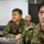 Three military personnel in Japanese Ground Self-Defense Force (JGSDF) camouflage uniforms are seated at a conference table in what appears to be a briefing or command room. A nameplate in Japanese characters reading '防空作戦調整所長' (Chief of Air Defense Operations Coordination Center) is visible on the table. The individuals appear attentive, likely participating in a military briefing or exercise.