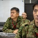 Three military personnel in Japanese Ground Self-Defense Force (JGSDF) camouflage uniforms are seated at a conference table in what appears to be a briefing or command room. A nameplate in Japanese characters reading '防空作戦調整所長' (Chief of Air Defense Operations Coordination Center) is visible on the table. The individuals appear attentive, likely participating in a military briefing or exercise.