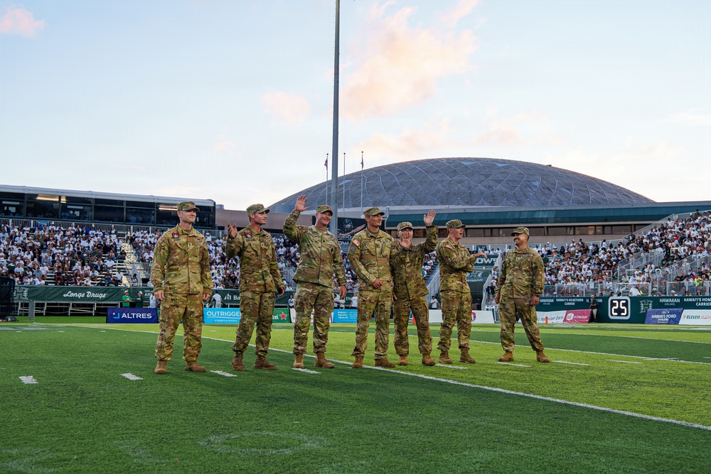 Seven military personnel in Army Combat Uniforms (ACU/OCP camouflage) are standing on a football field during what appears to be a re-enlistment or oath ceremony, with several raising their right hands. The event is taking place at a stadium in Hawaii, identifiable by the distinctive dome structure of the Stan Sheriff Center and sponsor banners including 'Outrigger' and 'Hawaiian Ford.' The stadium is filled with spectators in the background during what appears to be an evening game.