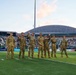 Seven military personnel in Army Combat Uniforms (ACU/OCP camouflage) are standing on a football field during what appears to be a re-enlistment or oath ceremony, with several raising their right hands. The event is taking place at a stadium in Hawaii, identifiable by the distinctive dome structure of the Stan Sheriff Center and sponsor banners including 'Outrigger' and 'Hawaiian Ford.' The stadium is filled with spectators in the background during what appears to be an evening game.