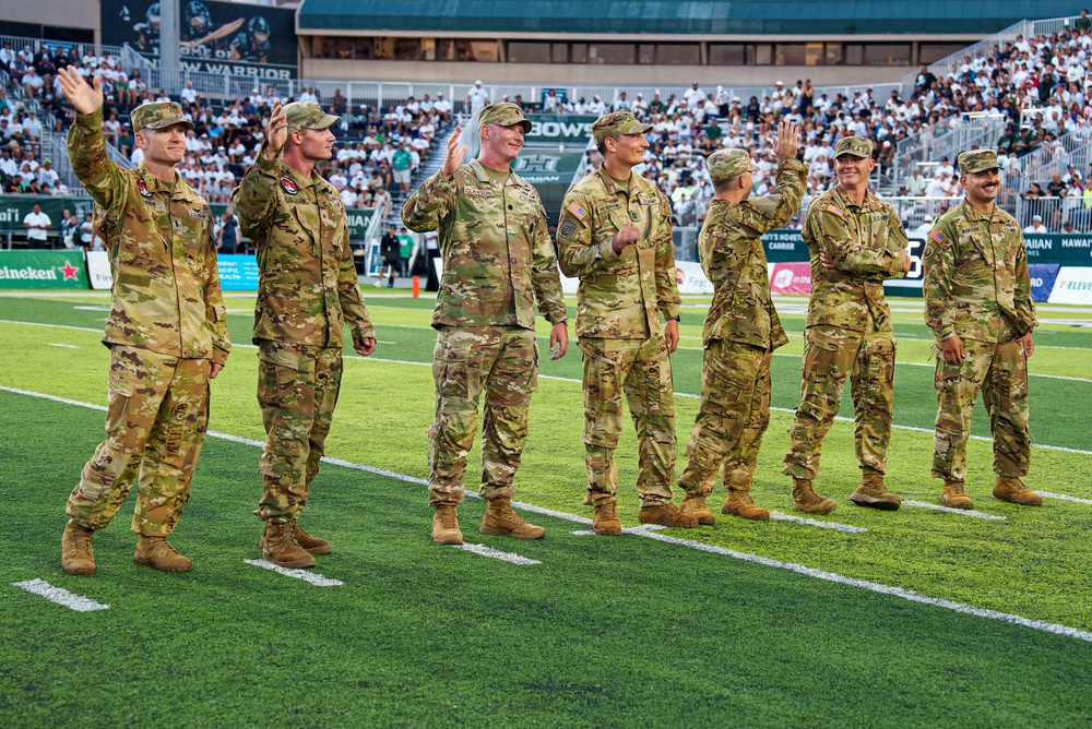 A group of approximately seven U.S. Army soldiers in OCP (Operational Camouflage Pattern) uniforms are standing on a football field during a ceremony, waving to the crowd. The setting appears to be the University of Hawaii's Clarence T.C. Ching Athletics Complex (Aloha Stadium area), identifiable by the 'Rainbow Warriors' branding and green turf. This appears to be a military appreciation or recognition ceremony during a college football game.