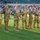 A group of approximately seven U.S. Army soldiers in OCP (Operational Camouflage Pattern) uniforms are standing on a football field during a ceremony, waving to the crowd. The setting appears to be the University of Hawaii's Clarence T.C. Ching Athletics Complex (Aloha Stadium area), identifiable by the 'Rainbow Warriors' branding and green turf. This appears to be a military appreciation or recognition ceremony during a college football game.