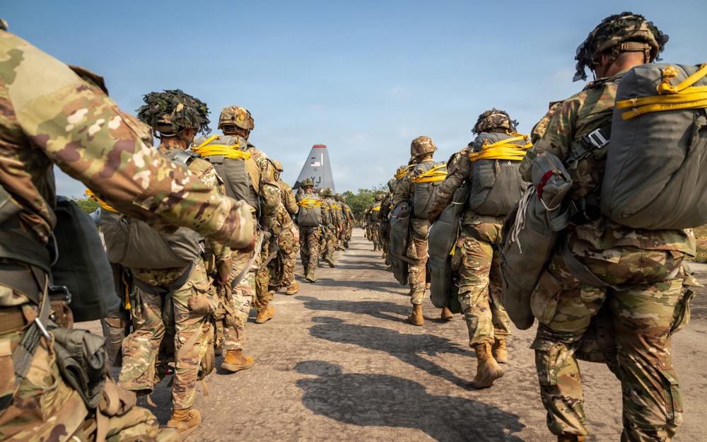 A column of military paratroopers in full combat gear and parachute equipment are marching toward a military transport aircraft, visible in the background. The soldiers are equipped with parachute packs, helmets, and tactical gear, suggesting they are preparing for an airborne operation or jump exercise. The image is taken from ground level looking down the center of two parallel lines of troops converging toward the aircraft.