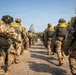 A column of military paratroopers in full combat gear and parachute equipment are marching toward a military transport aircraft, visible in the background. The soldiers are equipped with parachute packs, helmets, and tactical gear, suggesting they are preparing for an airborne operation or jump exercise. The image is taken from ground level looking down the center of two parallel lines of troops converging toward the aircraft.