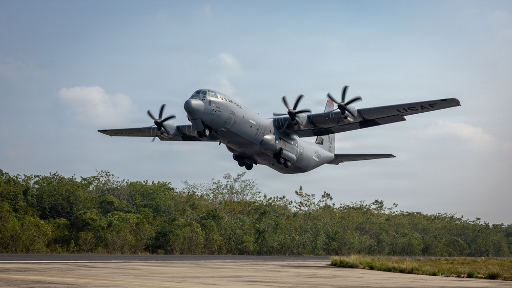 A U.S. Air Force C-130J Super Hercules military transport aircraft is captured at the moment of takeoff from a runway. The aircraft is painted in standard gray military livery with 'U.S. AIR FORCES' markings on the fuselage and 'USAF' on the tail. The setting appears to be a tropical or subtropical location, evidenced by the dense green tree line in the background.