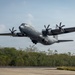 A U.S. Air Force C-130J Super Hercules military transport aircraft is captured at the moment of takeoff from a runway. The aircraft is painted in standard gray military livery with 'U.S. AIR FORCES' markings on the fuselage and 'USAF' on the tail. The setting appears to be a tropical or subtropical location, evidenced by the dense green tree line in the background.
