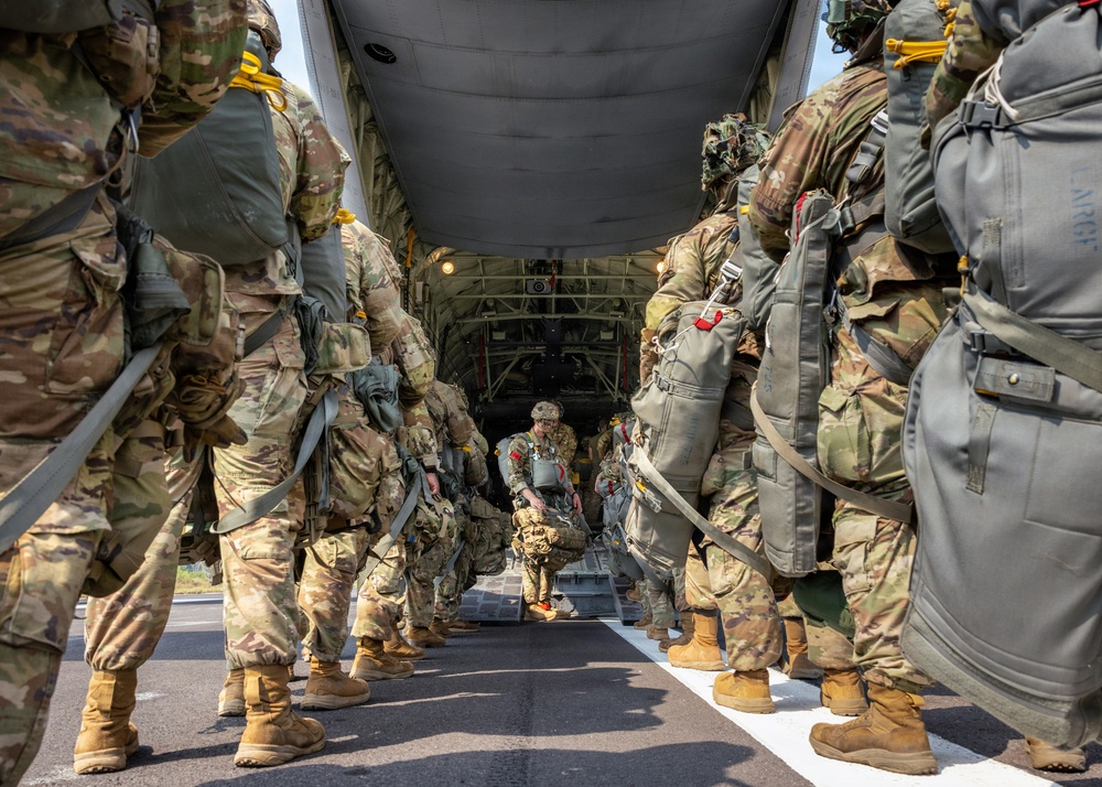 Military paratroopers in full combat gear are boarding or preparing to board a military transport aircraft, likely a C-130 Hercules, via its rear ramp. The soldiers are equipped with parachute packs, rucksacks, and full tactical gear in multicam/OCP camouflage pattern. The image is taken from ground level looking up the center aisle between two columns of troops toward the aircraft's open cargo ramp.