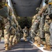 Military paratroopers in full combat gear are boarding or preparing to board a military transport aircraft, likely a C-130 Hercules, via its rear ramp. The soldiers are equipped with parachute packs, rucksacks, and full tactical gear in multicam/OCP camouflage pattern. The image is taken from ground level looking up the center aisle between two columns of troops toward the aircraft's open cargo ramp.