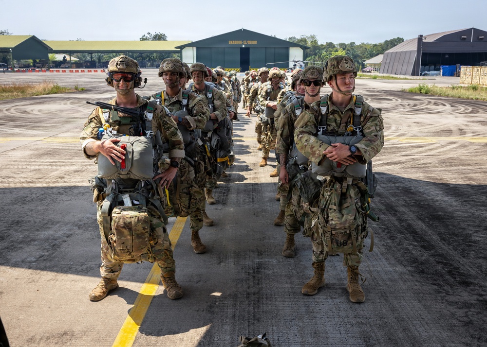 A group of military paratroopers in full jump gear are lined up in two columns on an airfield tarmac, apparently preparing for a parachute operation. The soldiers are wearing multicam camouflage uniforms and are equipped with parachute rigs, helmets, and tactical gear. A large aircraft hangar with text reading 'SQUADRON 12/SERON' is visible in the background.