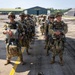 A group of military paratroopers in full jump gear are lined up in two columns on an airfield tarmac, apparently preparing for a parachute operation. The soldiers are wearing multicam camouflage uniforms and are equipped with parachute rigs, helmets, and tactical gear. A large aircraft hangar with text reading 'SQUADRON 12/SERON' is visible in the background.