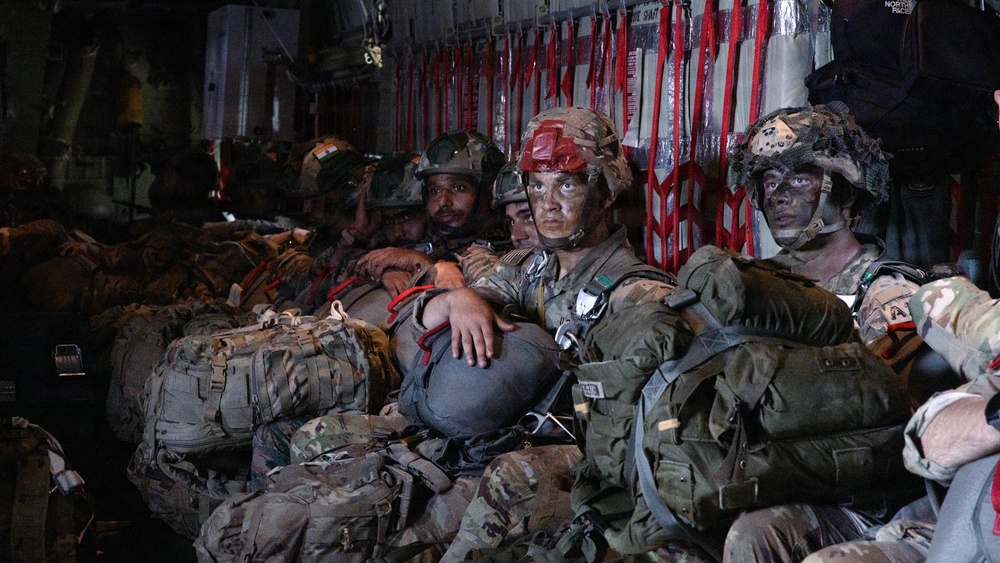 A group of military personnel in full combat gear are seated inside what appears to be a military transport aircraft, likely a C-130 or similar cargo plane. The soldiers are wearing camouflage uniforms, helmets with jump equipment, and have tactical face paint applied, suggesting preparation for a airborne/parachute operation. The interior of the aircraft is dimly lit with red safety straps and netting visible along the fuselage walls.