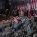 A group of military personnel in full combat gear are seated inside what appears to be a military transport aircraft, likely a C-130 or similar cargo plane. The soldiers are wearing camouflage uniforms, helmets with jump equipment, and have tactical face paint applied, suggesting preparation for a airborne/parachute operation. The interior of the aircraft is dimly lit with red safety straps and netting visible along the fuselage walls.