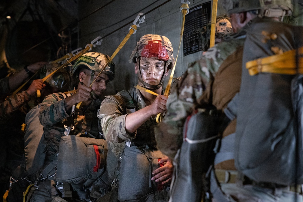 Military paratroopers are seated inside a military transport aircraft, preparing for a static-line jump operation. The soldiers are wearing full combat gear including helmets, parachute rigs, and equipment bundles, and are holding their static lines. At least two distinct military uniforms are visible, suggesting a joint or multinational operation.