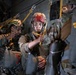 Military paratroopers are seated inside a military transport aircraft, preparing for a static-line jump operation. The soldiers are wearing full combat gear including helmets, parachute rigs, and equipment bundles, and are holding their static lines. At least two distinct military uniforms are visible, suggesting a joint or multinational operation.
