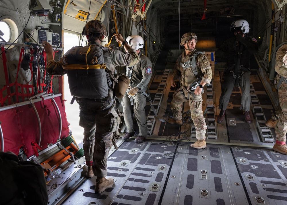Military personnel are positioned inside the open rear cargo ramp of a large military transport aircraft, appearing to be preparing for a parachute jump operation. Several soldiers in full combat gear and parachute equipment are visible, with one crew member wearing a distinctive white flight helmet. The aircraft interior shows standard cargo configuration with red safety netting, tie-down rails, and equipment secured along the walls.