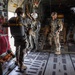 Military personnel are positioned inside the open rear cargo ramp of a large military transport aircraft, appearing to be preparing for a parachute jump operation. Several soldiers in full combat gear and parachute equipment are visible, with one crew member wearing a distinctive white flight helmet. The aircraft interior shows standard cargo configuration with red safety netting, tie-down rails, and equipment secured along the walls.