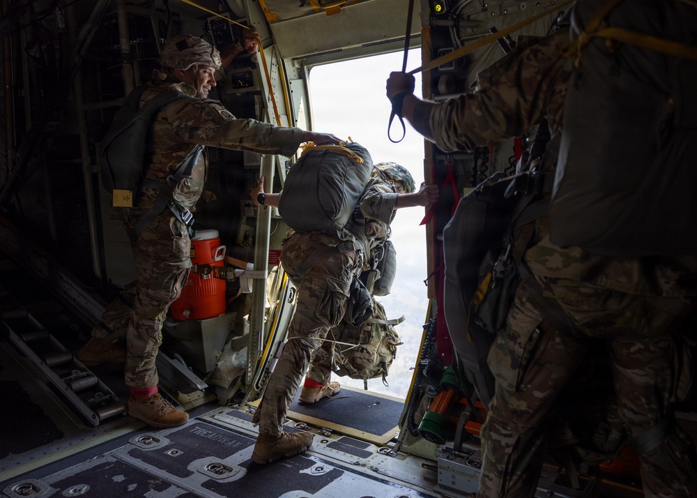 Military personnel are conducting a static-line parachute jump from what appears to be a C-130 Hercules transport aircraft. A soldier carrying a large rucksack and parachute pack is in the process of exiting through the open side door of the aircraft, while a jumpmaster assists and monitors the jump. The interior of the aircraft is visible, including safety equipment and a static line attached overhead.