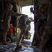 Military personnel are conducting a static-line parachute jump from what appears to be a C-130 Hercules transport aircraft. A soldier carrying a large rucksack and parachute pack is in the process of exiting through the open side door of the aircraft, while a jumpmaster assists and monitors the jump. The interior of the aircraft is visible, including safety equipment and a static line attached overhead.