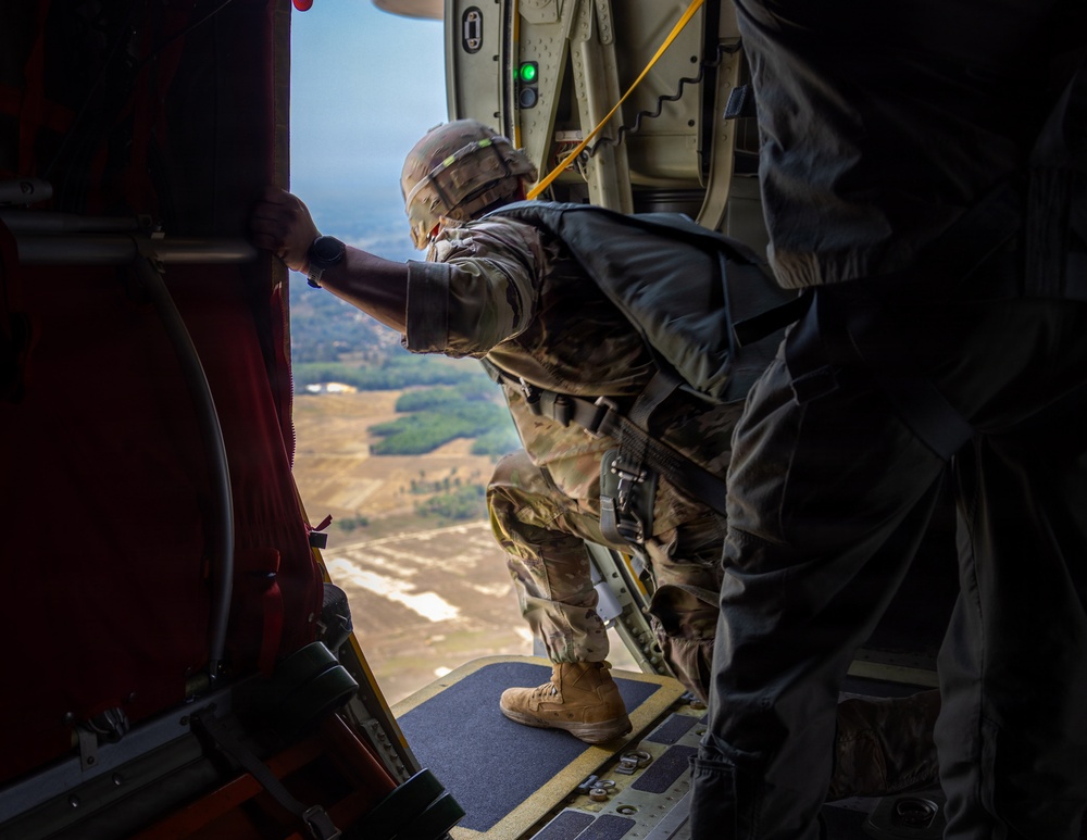 A military paratrooper in full combat gear and camouflage uniform is positioned at the open door of a military aircraft, leaning out to observe the terrain below. The individual appears to be preparing for or conducting a static-line parachute jump, evidenced by the yellow static line visible overhead. The landscape visible below shows open fields and wooded areas at significant altitude.