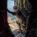 A military paratrooper in full combat gear and camouflage uniform is positioned at the open door of a military aircraft, leaning out to observe the terrain below. The individual appears to be preparing for or conducting a static-line parachute jump, evidenced by the yellow static line visible overhead. The landscape visible below shows open fields and wooded areas at significant altitude.