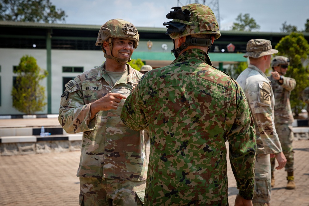 The image shows military personnel from what appear to be different nations engaged in conversation at an outdoor military installation. A U.S. Army soldier identified by the name tape 'STEPPE' wearing Operational Camouflage Pattern (OCP) uniform and helmet is speaking with a soldier in a different camouflage pattern, likely Japanese Ground Self-Defense Force based on the distinctive uniform. Additional U.S. soldiers are visible in the background near a building with green trim.