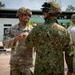 The image shows military personnel from what appear to be different nations engaged in conversation at an outdoor military installation. A U.S. Army soldier identified by the name tape 'STEPPE' wearing Operational Camouflage Pattern (OCP) uniform and helmet is speaking with a soldier in a different camouflage pattern, likely Japanese Ground Self-Defense Force based on the distinctive uniform. Additional U.S. soldiers are visible in the background near a building with green trim.