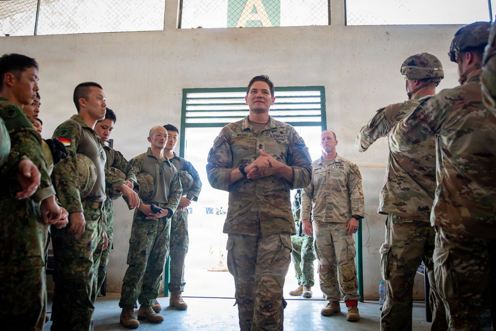 A U.S. Army soldier, identified by name tape as 'WOOD,' stands at the center of a group of multinational military personnel inside what appears to be a training facility or hangar. The gathering includes soldiers in different camouflage patterns, suggesting a joint or combined military exercise. The central figure appears to be addressing or briefing the assembled group.