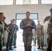 A U.S. Army soldier, identified by name tape as 'WOOD,' stands at the center of a group of multinational military personnel inside what appears to be a training facility or hangar. The gathering includes soldiers in different camouflage patterns, suggesting a joint or combined military exercise. The central figure appears to be addressing or briefing the assembled group.