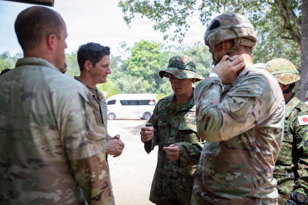 A group of military personnel from multiple nations are engaged in a discussion in an outdoor setting. The group includes soldiers wearing US Army multicam uniforms and Japanese Ground Self-Defense Force uniforms, suggesting a joint military exercise or coordination meeting. The setting appears to be a tropical or subtropical environment based on the vegetation visible in the background.