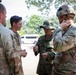 A group of military personnel from multiple nations are engaged in a discussion in an outdoor setting. The group includes soldiers wearing US Army multicam uniforms and Japanese Ground Self-Defense Force uniforms, suggesting a joint military exercise or coordination meeting. The setting appears to be a tropical or subtropical environment based on the vegetation visible in the background.