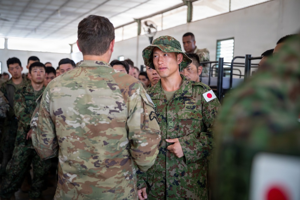 The image shows a military interaction between a U.S. Army soldier in OCP (Operational Camouflage Pattern) uniform and a Japan Ground Self-Defense Force (JGSDF) soldier wearing a distinctive Japanese camouflage uniform with a Japanese flag patch and boonie hat. The setting appears to be a barracks or indoor military facility, with a large group of Japanese soldiers visible in the background. This appears to be a joint military exercise or training event between U.S. and Japanese forces.