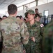 The image shows a military interaction between a U.S. Army soldier in OCP (Operational Camouflage Pattern) uniform and a Japan Ground Self-Defense Force (JGSDF) soldier wearing a distinctive Japanese camouflage uniform with a Japanese flag patch and boonie hat. The setting appears to be a barracks or indoor military facility, with a large group of Japanese soldiers visible in the background. This appears to be a joint military exercise or training event between U.S. and Japanese forces.