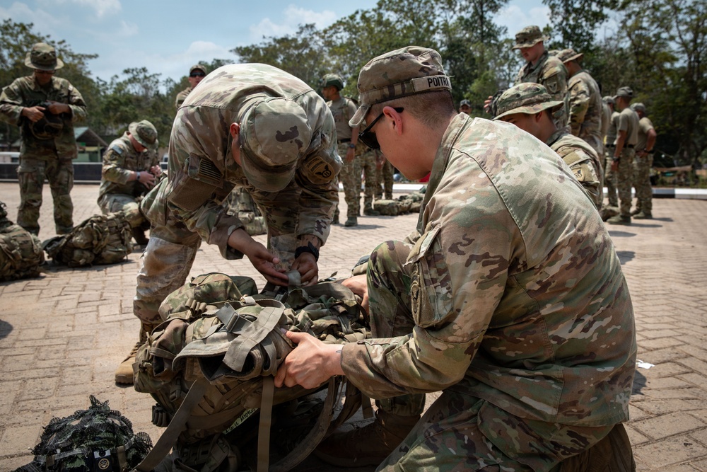 Military personnel in MultiCam/OCP camouflage uniforms are engaged in equipment preparation or inspection on a paved outdoor area. Two soldiers in the foreground are working together on a large tactical rucksack or pack assembly. The scene appears to be a training or pre-mission equipment check with multiple soldiers performing similar tasks in the background.