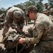 Military personnel in MultiCam/OCP camouflage uniforms are engaged in equipment preparation or inspection on a paved outdoor area. Two soldiers in the foreground are working together on a large tactical rucksack or pack assembly. The scene appears to be a training or pre-mission equipment check with multiple soldiers performing similar tasks in the background.