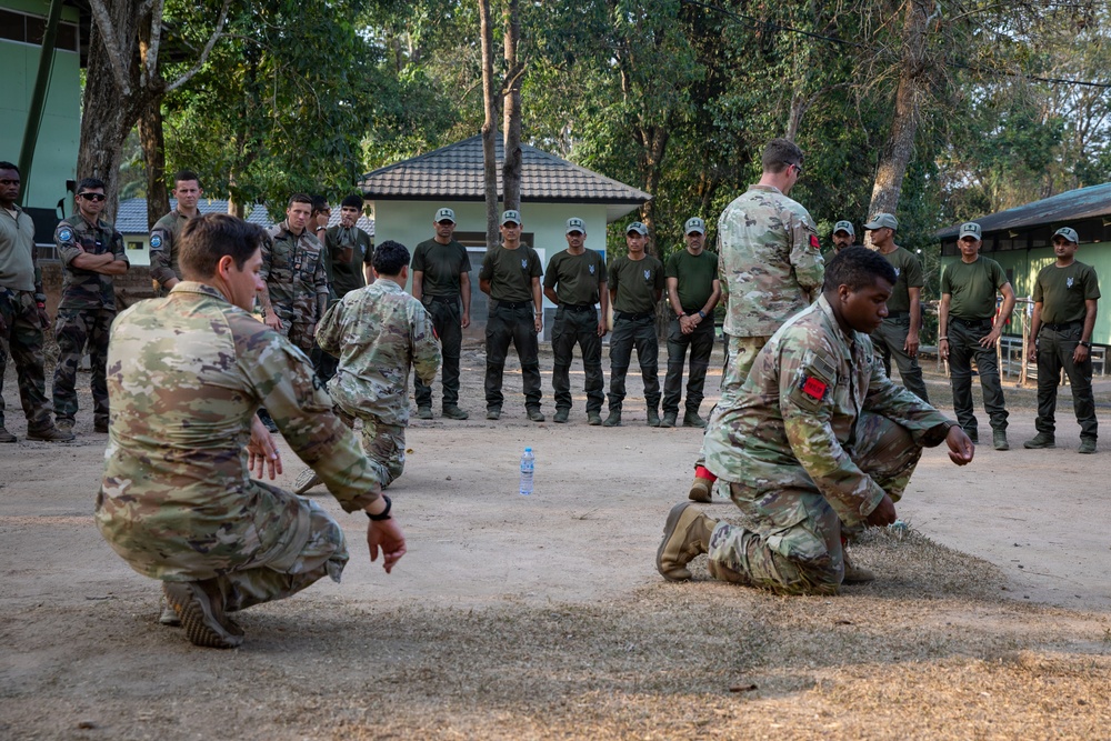 A military training exercise is taking place outdoors at what appears to be a multinational military installation, likely in Southeast Asia based on the architecture and vegetation. Several soldiers in US Army MultiCam/OCP uniforms are demonstrating or practicing ground-level techniques while a larger group of observers in olive drab uniforms and caps watch from a semicircle. The setting suggests a joint training event or military exercise involving multiple nations.