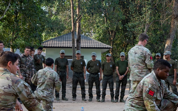 A military training exercise is taking place outdoors at what appears to be a multinational military installation, likely in Southeast Asia based on the architecture and vegetation. Several soldiers in US Army MultiCam/OCP uniforms are demonstrating or practicing ground-level techniques while a larger group of observers in olive drab uniforms and caps watch from a semicircle. The setting suggests a joint training event or military exercise involving multiple nations.