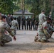 A military training exercise is taking place outdoors at what appears to be a multinational military installation, likely in Southeast Asia based on the architecture and vegetation. Several soldiers in US Army MultiCam/OCP uniforms are demonstrating or practicing ground-level techniques while a larger group of observers in olive drab uniforms and caps watch from a semicircle. The setting suggests a joint training event or military exercise involving multiple nations.