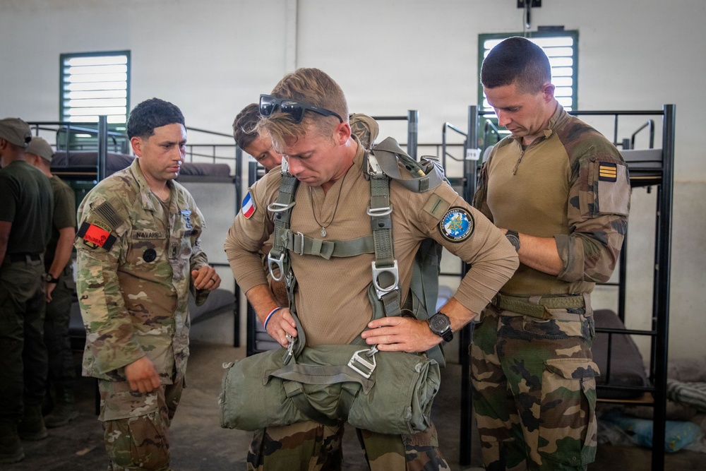 Military personnel from what appears to be a joint US-French operation are gathered inside a barracks or staging area. A central figure wearing a French military uniform with a French flag patch is being fitted or inspected with a parachute harness. Other soldiers, including one in US Army multicam uniform with a 'NAVARRO' name tape, observe and assist.