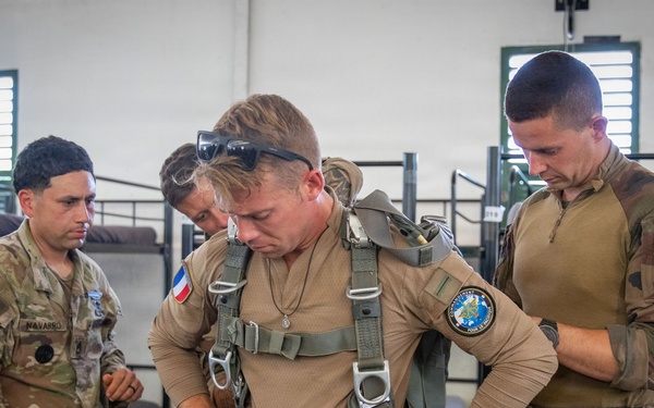 Military personnel from what appears to be a joint US-French operation are gathered inside a barracks or staging area. A central figure wearing a French military uniform with a French flag patch is being fitted or inspected with a parachute harness. Other soldiers, including one in US Army multicam uniform with a 'NAVARRO' name tape, observe and assist.