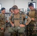 Military personnel from what appears to be a joint US-French operation are gathered inside a barracks or staging area. A central figure wearing a French military uniform with a French flag patch is being fitted or inspected with a parachute harness. Other soldiers, including one in US Army multicam uniform with a 'NAVARRO' name tape, observe and assist.