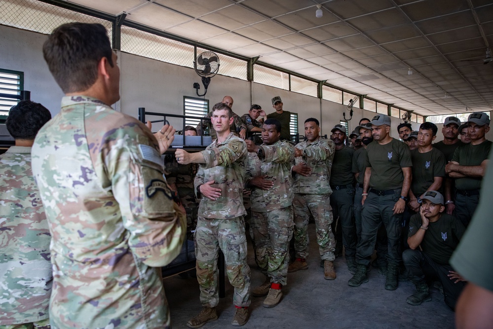 A military training session is taking place inside an open-sided covered facility, where a U.S. Army soldier in multicam uniform is demonstrating a technique to a mixed group of personnel. Several U.S. soldiers in OCP/multicam uniforms are actively participating in what appears to be a hands-on combatives or detainee handling demonstration. A larger group of local or partner-nation military/security forces in olive drab uniforms observe the instruction.