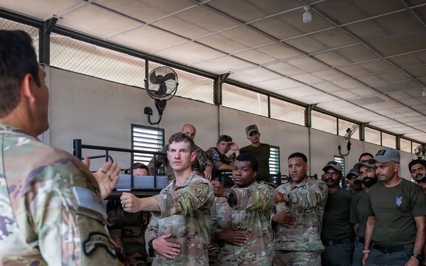 A military training session is taking place inside an open-sided covered facility, where a U.S. Army soldier in multicam uniform is demonstrating a technique to a mixed group of personnel. Several U.S. soldiers in OCP/multicam uniforms are actively participating in what appears to be a hands-on combatives or detainee handling demonstration. A larger group of local or partner-nation military/security forces in olive drab uniforms observe the instruction.