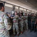 A military training session is taking place inside an open-sided covered facility, where a U.S. Army soldier in multicam uniform is demonstrating a technique to a mixed group of personnel. Several U.S. soldiers in OCP/multicam uniforms are actively participating in what appears to be a hands-on combatives or detainee handling demonstration. A larger group of local or partner-nation military/security forces in olive drab uniforms observe the instruction.