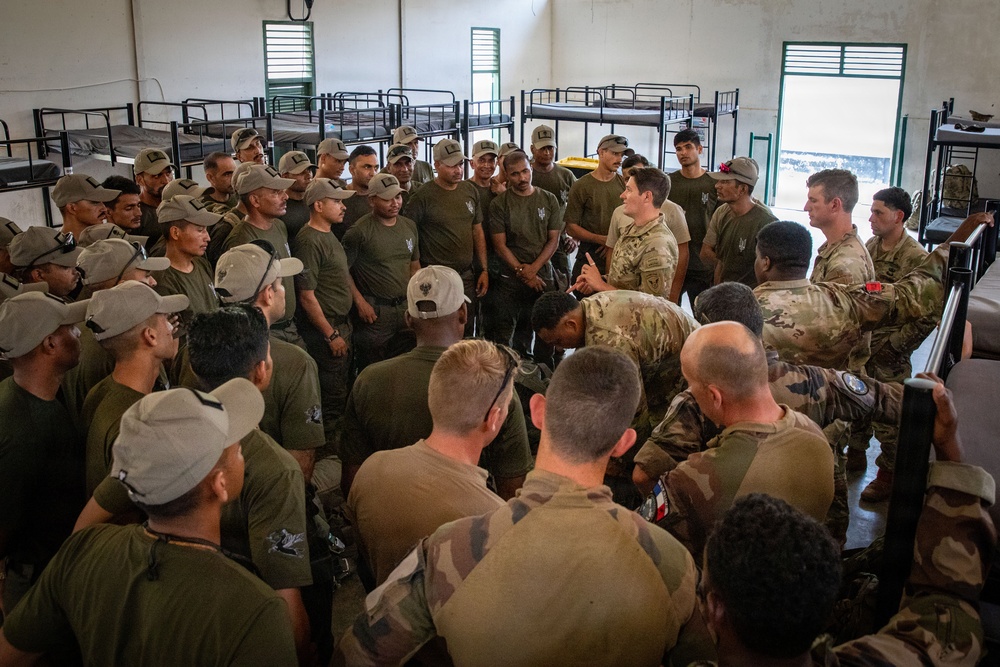 A large group of military personnel gathered inside a barracks room with bunk beds visible in the background. The group appears to include soldiers in different uniforms — olive/dark green uniforms with caps and camouflage pattern uniforms — suggesting a multinational or joint military exercise or briefing. One individual in camouflage appears to be addressing or demonstrating something to the assembled group.