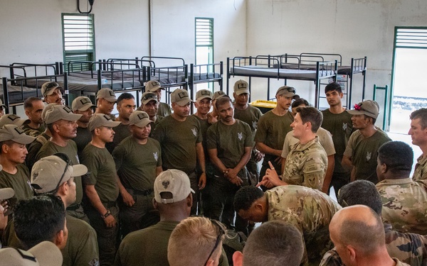 A large group of military personnel gathered inside a barracks room with bunk beds visible in the background. The group appears to include soldiers in different uniforms — olive/dark green uniforms with caps and camouflage pattern uniforms — suggesting a multinational or joint military exercise or briefing. One individual in camouflage appears to be addressing or demonstrating something to the assembled group.