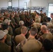 A large group of military personnel gathered inside a barracks room with bunk beds visible in the background. The group appears to include soldiers in different uniforms — olive/dark green uniforms with caps and camouflage pattern uniforms — suggesting a multinational or joint military exercise or briefing. One individual in camouflage appears to be addressing or demonstrating something to the assembled group.