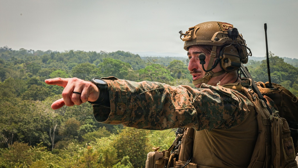 A military service member in full combat gear is pointing toward a distant location while overlooking a dense forested landscape. The soldier is equipped with advanced tactical equipment including a helmet-mounted communication system and a radio antenna. The setting appears to be an elevated position above a tropical or subtropical forest environment.