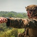 A military service member in full combat gear is pointing toward a distant location while overlooking a dense forested landscape. The soldier is equipped with advanced tactical equipment including a helmet-mounted communication system and a radio antenna. The setting appears to be an elevated position above a tropical or subtropical forest environment.