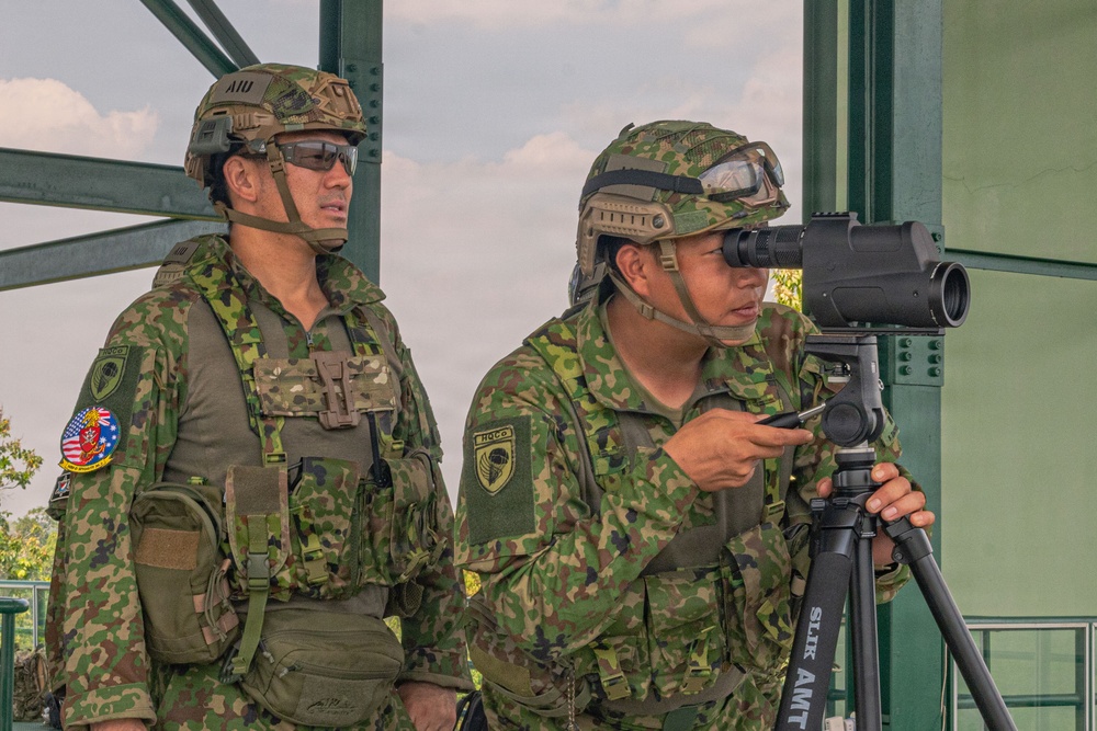 Two military personnel in camouflage uniforms are positioned at what appears to be an observation post or elevated platform. One soldier is looking through a spotting scope mounted on a tripod, while the other stands watch. Both wear tactical helmets with goggles and display unit patches on their uniforms.